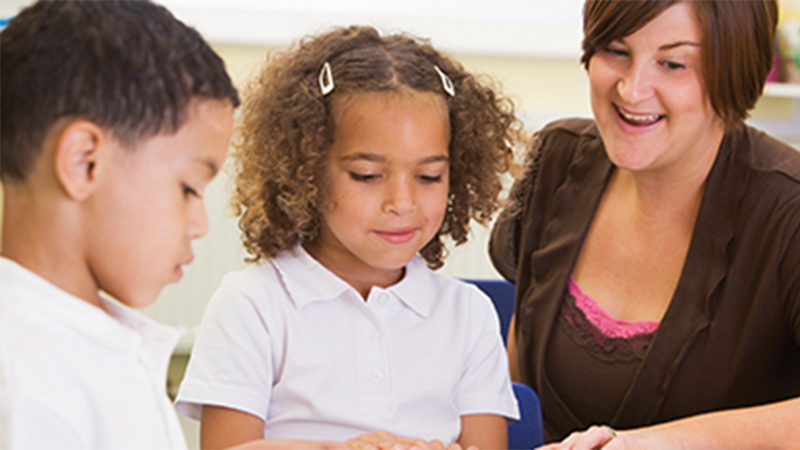 Two elementary schoolers being assisted by a teacher while reading a book