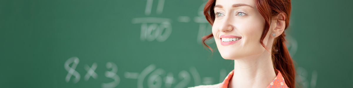 A smiling red-haired woman stands in front of a green chalkboard with math equations written on it.