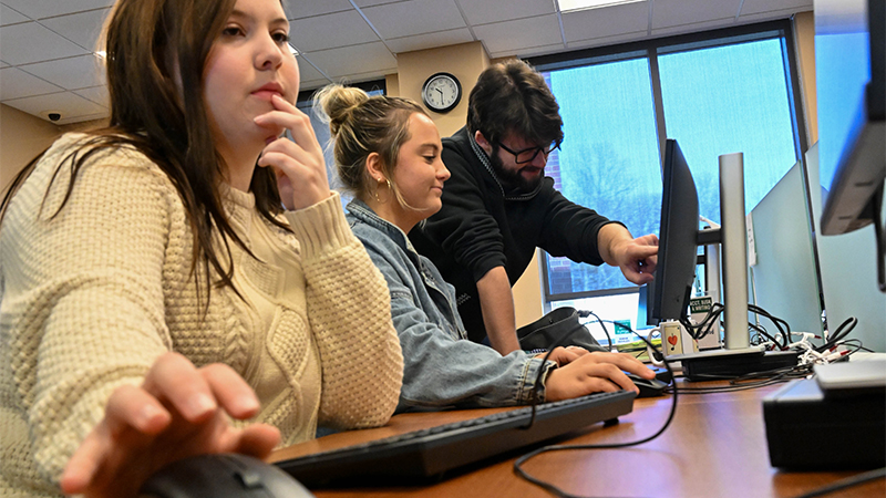 Three students engaged using computers. The female student, sits at a desk using a mouse, while another female student listens attentively to the man pointing at the screen.