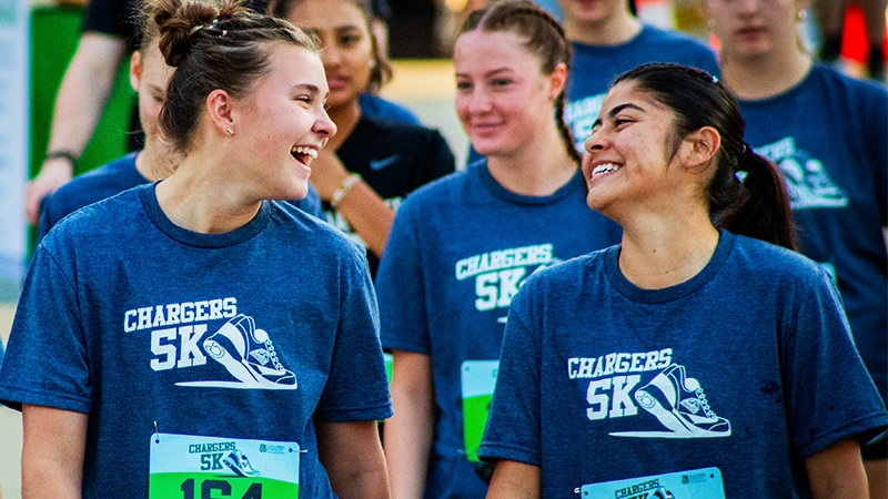 Two young women wearing navy blue "Chargers 5K" t-shirts smile and laughing, with race numbers pinned to their chests.