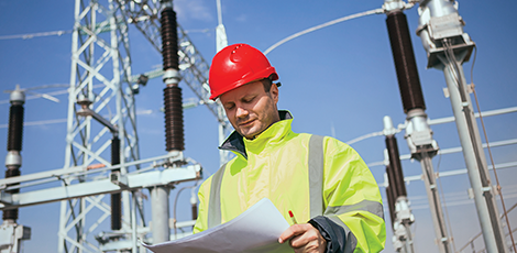 A man in a hard hat and a yellow reflective jacket standing in the center of an electric substation