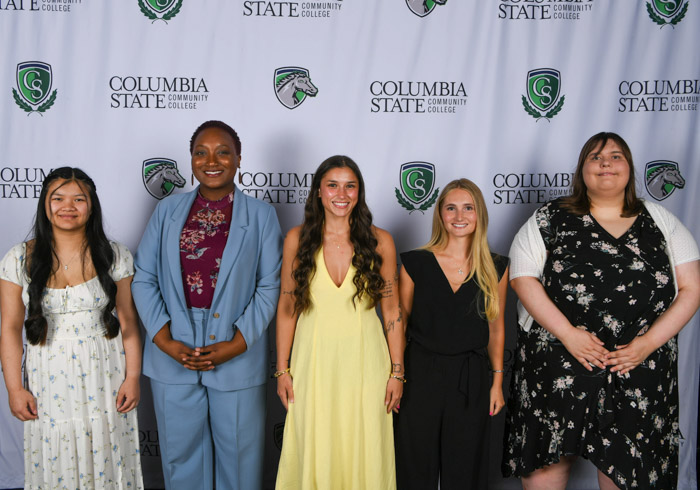 A group of five women stand in front of a background.