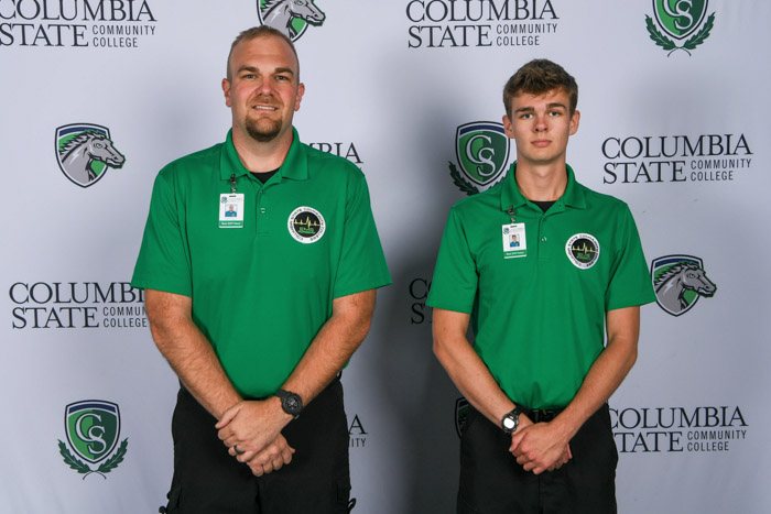 Two men stand in front of a white background.